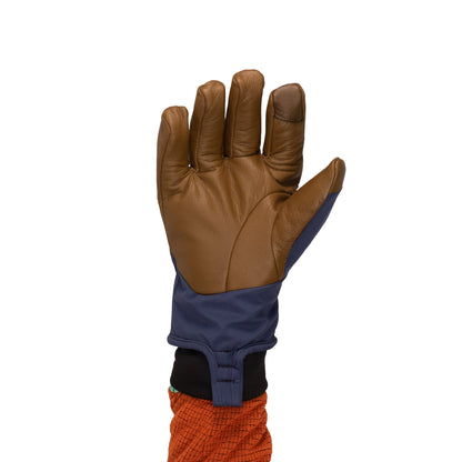 The Loaf Glove by Mainers is recognized for its warmth, showcasing a leather design complemented by a blue fabric sleeve covering the top side of the hand, while an orange jacket sleeve subtly emerges from underneath.