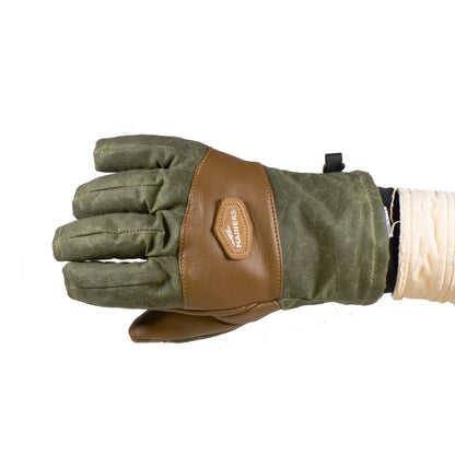 A green and brown Mainers Ragged MT Glove, featuring a "AMERICAS" leather patch, is displayed horizontally against a white background. Constructed from waxed canvas and leather, this versatile winter glove is perfect for various uses.